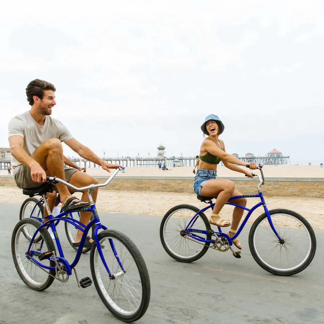 A couple of friends ride bicycles on the beach path, with a historical background shot of Ruby's on the pier.