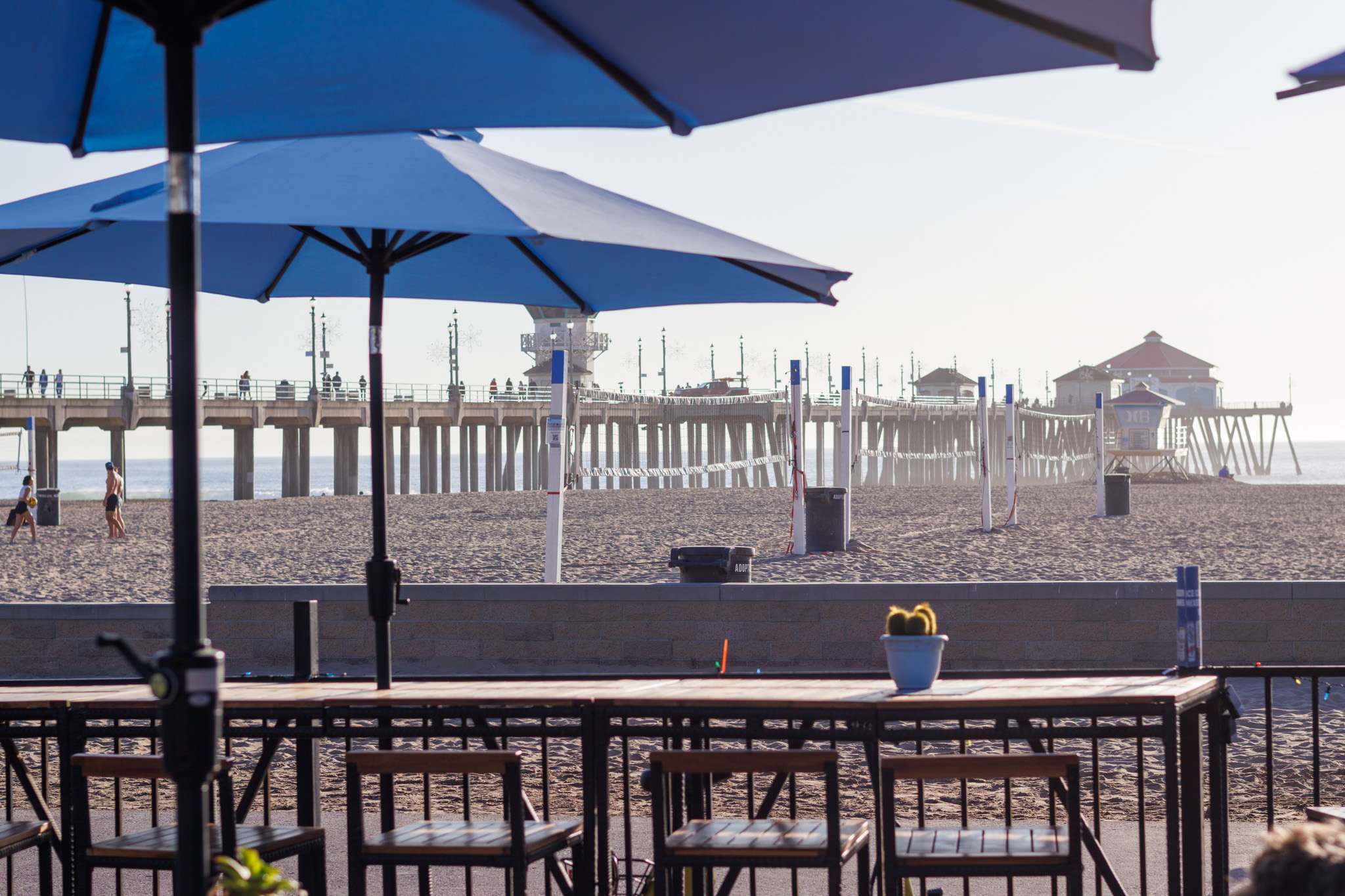 Outdoor seating area with blue umbrellas and tables near a pier on a sunny day in Huntington Beach California